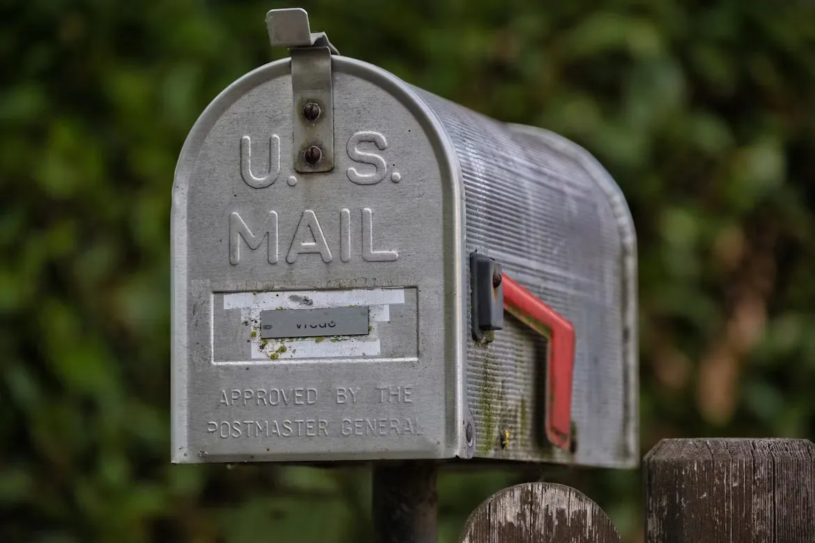 A weathered metal mailbox marked U.S. Mail with a red flag beside a wooden fence.