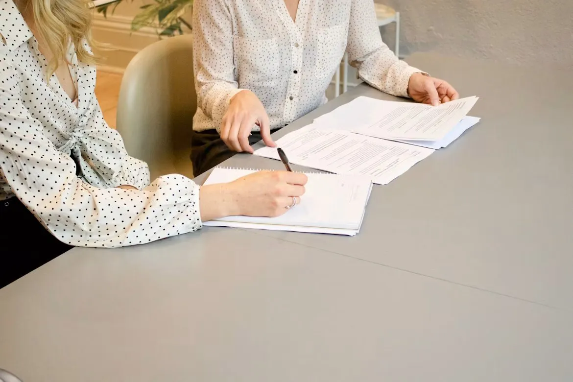 Two colleagues review printed documents while one writes notes at a meeting table.