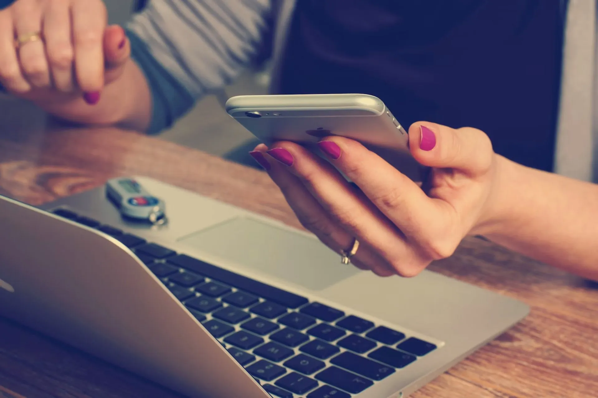 A person checks a smartphone beside an open laptop on a wooden desk.