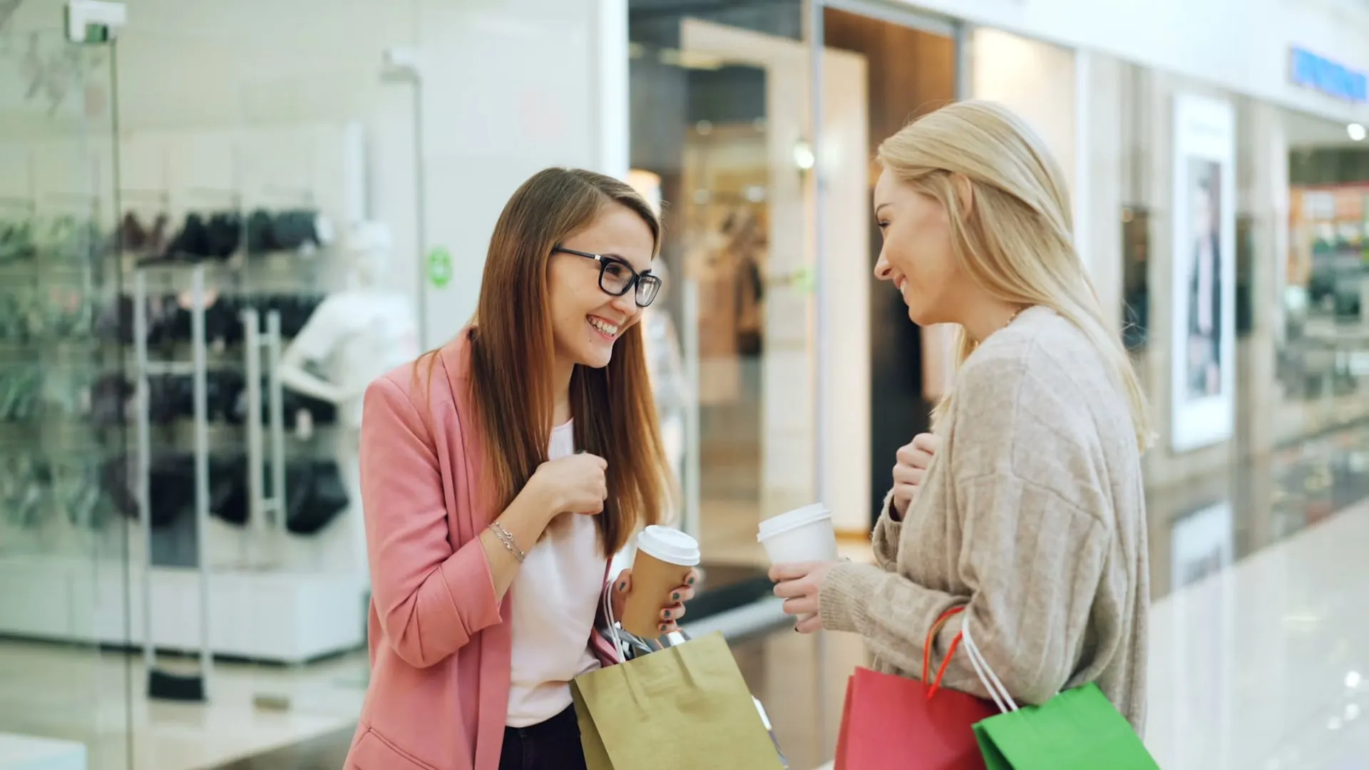 Two friends talk while holding shopping bags and coffee in a shopping center.