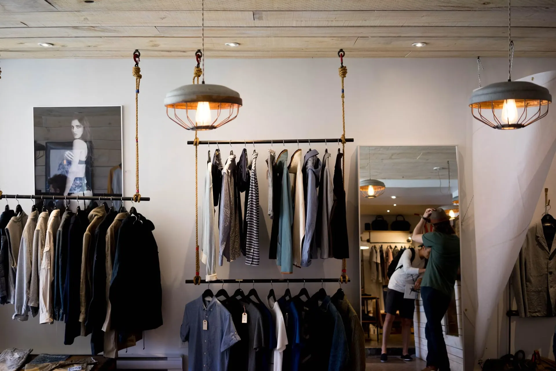 A clothing boutique with suspended display racks, pendant lights, and a large mirror.
