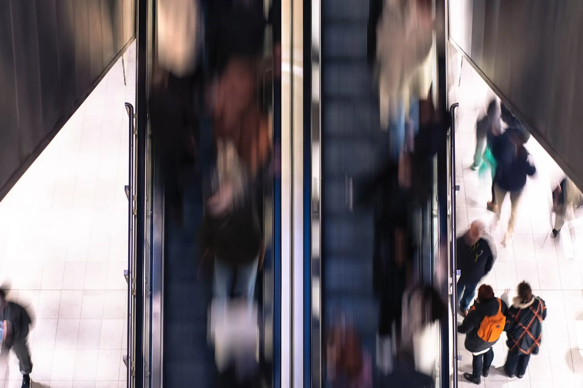 Motion-blurred shoppers move along escalators and walkways in a busy shopping center.