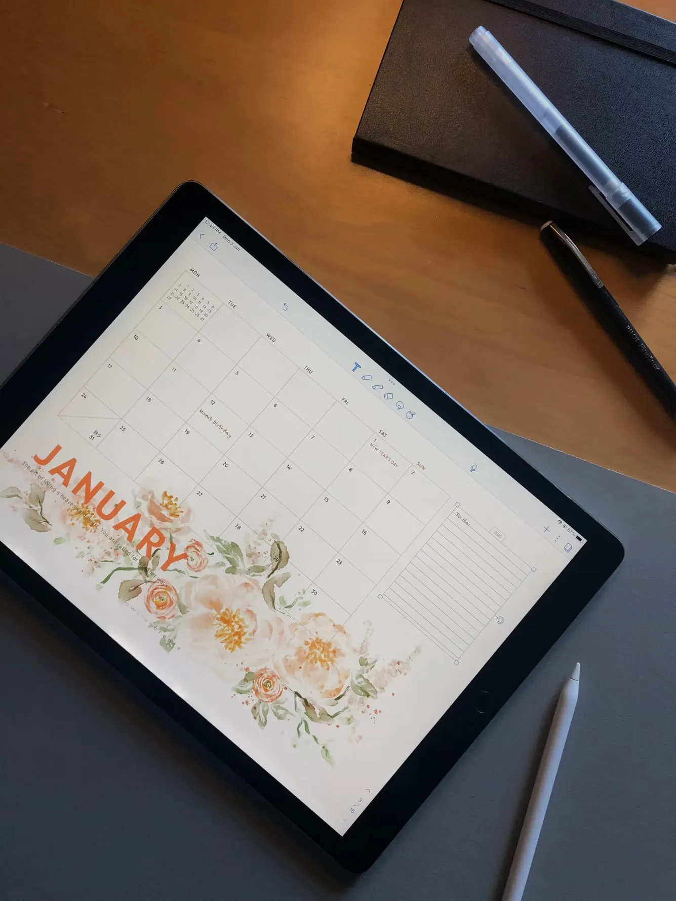A monthly calendar displayed on a tablet beside notebooks and pens.