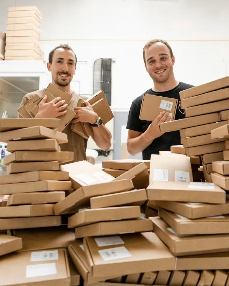 Two workers stand at a packing table surrounded by prepared cardboard parcels.